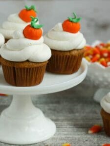 Pumpkin Cupcakes with Whipped Cream Frosting on a cake stand and decorated with fondant pumpkins and served with a bowl of candy corn.
