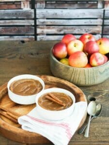 Homemade Applesauce served in a white bowl garnished with cinnamon sticks served next to a wooden bowl of red apples.