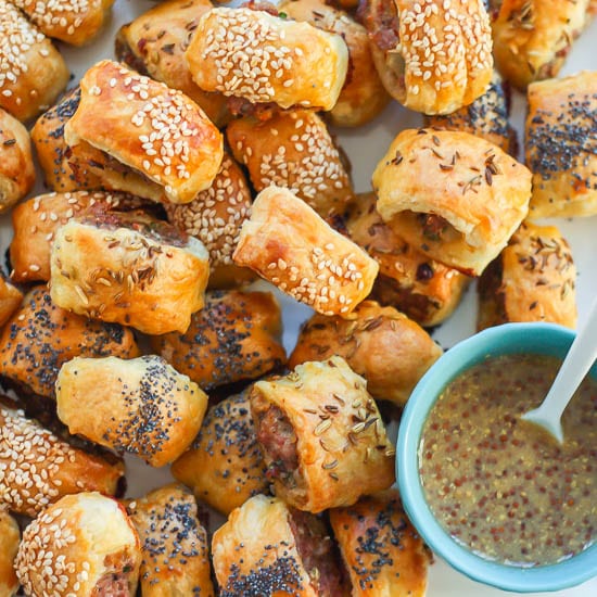 A white square plate is piled with Sausage Rolls that are golden brown, sprinkled with loads of seeds and have a little cup of mustard for dipping nearby.