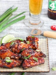 Crispy Asian Chicken Wings served on a square plate, sprinkled with sesame seeds, scallion slices, and lime wedges with a beer and bottle opener in the background.