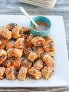 A white square plate is piled with Sausage Rolls that are golden brown, sprinkled with loads of seeds and have a little cup of mustard for dipping nearby.