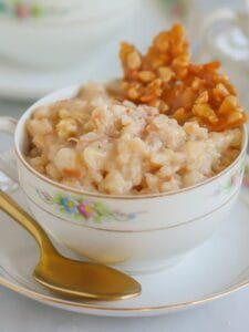 Coconut Rice Pudding with Coconut Caramel Brittle served in a pretty china cup on a saucer with a gold spoon alongside.