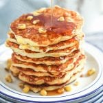 A stack of Corn Pancakes piled on a stack of white an blue plates with corn kernels scattered on top and big drizzle of syrup being poured over.