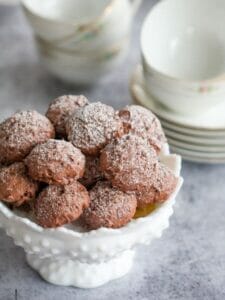 Chocolate Meringue Cookies dusted with confectioners sugar piled in a pretty white bowl with cups and saucers stacked in the background.