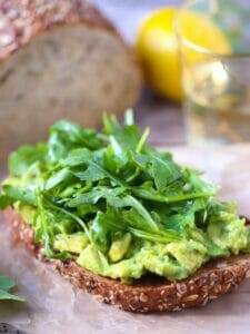 Lemony Avocado Toast with Arugula with a lemon, loaf of multigrain bread and a glass of white wine in the background.