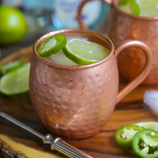 Close up shot of a Mexican Mule served in a copper mug garnished with lime and jalapeño slices placed on a wood board with another mug in the background.