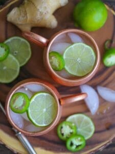 Overhead shot of two Mexican Mules in copper mugs garnished with lime and jalapeño slices on a wood board with ginger, lime, and jalapeños scattered around.