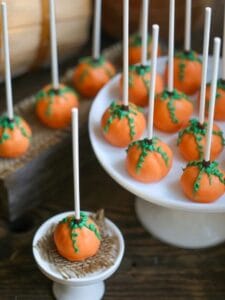 A festive display of pumpkin cake pops on a cake stand, cupcake stand with sticks facing up and decorative green vines on the orange cake pops.