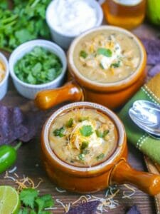 Two bowls of white chicken chili topped with sour cream, cheese, and cilantro served with beer, toppings, and blue corn chips.