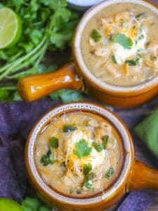 Two bowls of white chicken chili topped with sour cream, cheese, and cilantro served with toppings and blue corn chips.