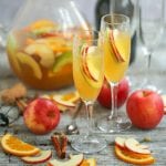 Sparkling Autumn Sangria served in two champagne flutes with apple slice garnishes with a pitcher of sangria in the background and fruit garnishes scattered on the table.