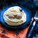 A Spooky Spaghetti Mummy on top of a black plate and stack of orange napkins with plastic spiders scattered around for decoration.