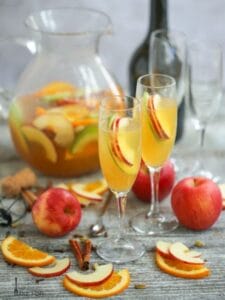 Sparkling Autumn Sangria served in two champagne flutes with apple slice garnishes with a pitcher of sangria in the background and fruit garnishes scattered on the table.