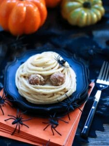 A Spooky Spaghetti Mummy on top of a black plate and stack of orange napkins with plastic spiders and pumpkins scattered around for decoration.