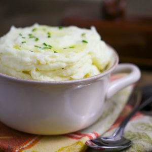 Creamy Mashed Potatoes topped with melted butter and chives in a bowl with a serving spoon on the side, on top of a folded fall napkin.
