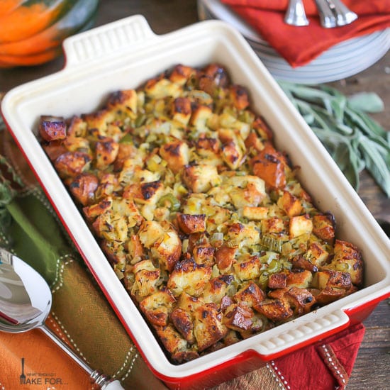 Pancetta and Leek Stuffing baked in a rectangle pan with crispy, buttery edges. The pan is placed on a autumn napkin, with a serving spoon and herbs alongside.
