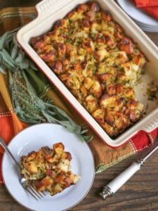 Pancetta and Leek Stuffing baked in a rectangle pan with crispy, buttery edges. A serving is scooped onto a plate with fresh herbs scattered around.