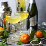 Sparkling Orange and St-Germain Cocktails are served in coupe glasses with orange peel twists, with clementines, silver noisemakers, and bar accessories scattered around.