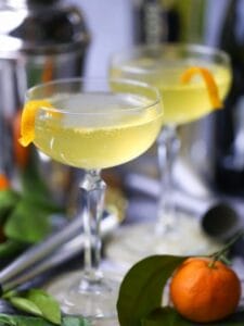 Sparkling Orange and St-Germain Cocktails are served in coupe glasses with orange peel twists, with clementines, silver noisemakers, and bar accessories scattered around.