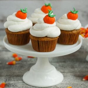 Pumpkin Cupcakes with Whipped Cream Frosting on a cake stand and decorated with fondant pumpkins and served with a bowl of candy corn.
