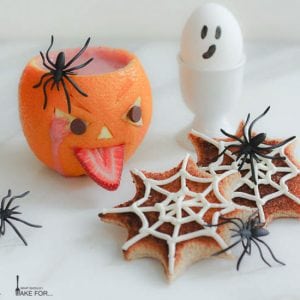 A spooky Halloween breakfast display: a decorated orange jack o'lantern filled with a strawberry smoothie, spiderweb decorated toast, and a ghostly hard boiled egg.