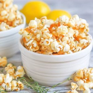 A close up of a bowl of Lemon Thyme Caramel Corn with lemons in the background and fresh thyme scattered in front.