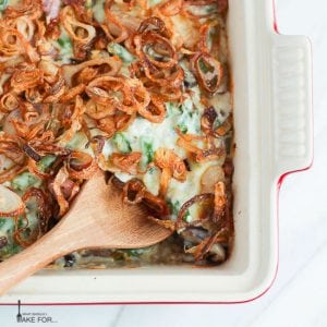 Creamy green bean casserole topped with crispy fried shallots in a rectangle baking dish with a wooden spoon scooping out a serving.