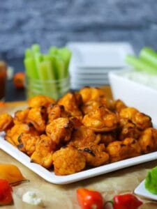 Buffalo Cauliflower Bites with Blue Cheese Dip served on a white square platter with celery stalks and a beer in the background.