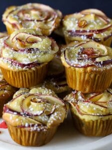 Apple Rose Cupcakes on a cake stand dusted with confectioners sugar and little candy hearts scattered on the plate with a glass of milk in the background..