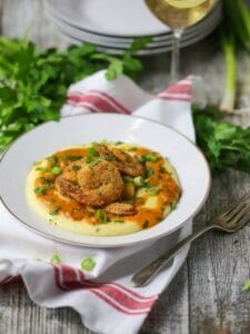 Italian Style Shrimp and Grits, three cornmeal crusted shrimp sit atop creamy polenta and tomato sauce, sprinkled with parsley and scallions on a white towel with red stripes, served with a glass of white wine.