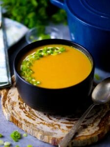 A bowl of carrot ginger soup garnished with scallions on a wood coaster with a blue pot, herbs and spoons in the background.