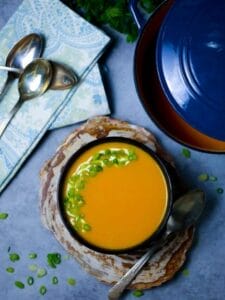 Overhead shot of a bowl of carrot ginger soup garnished with scallions on a wood coaster with a blue pot, herbs and spoons in the background.