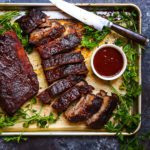 Baby Back Ribs with Bourbon BBQ Sauce on a sheet tray with herbs, sauce and cut with a knife.