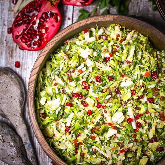 Brussels Sprouts and Pomegranate Salad served in a round wood bowl with pomegranate and apple slivers scattered around on the table.