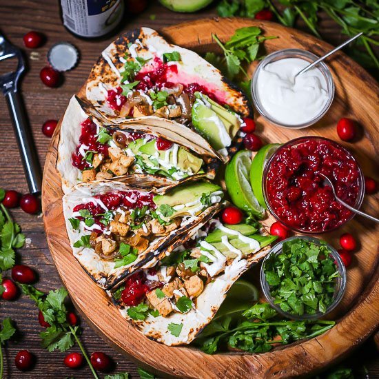 Four Leftover Turkey Tacos on a round wooden tray with cranberry chutney, minced cilantro and sour cream in small bowls with whole cranberries and cilantro scattered around.