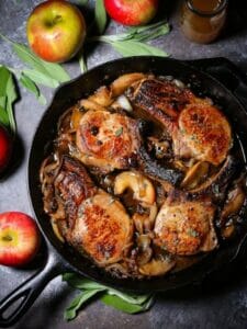 Apple cider pork chops in a cast iron skillet surrounded by apples and sage.