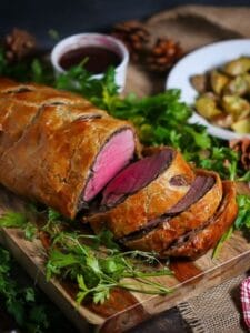 Beef Wellington with Red Wine Sauce sliced on a cutting board garnished with fresh parsley and a bowl of roasted potatoes in the background.