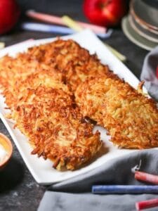 Celery Root Latkes on a white serving tray with apples, sour cream, apples, sauce, and menorah candles scattered around.