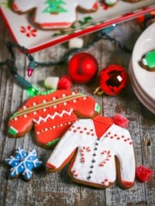 Two Ugly Sweater Gingerbread Cookies are laying on a wood table with gum drops, a mini snowflake cookie and red bulbs scattered around.