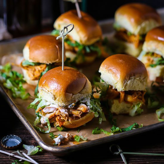 Buffalo Chicken Sliders skewered with knotted toothpicks on a sheet tray with beer in the background and a bottle cap on the table.