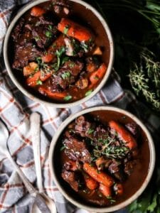 Overhead shot of two bowls of Beef Bourguignon filled with tender beef and carrots, placed on a plaid tablecloth with herbs in the background.