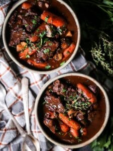 Overhead shot of two bowls of Beef Bourguignon filled with tender beef and carrots, placed on a plaid tablecloth with herbs in the background.
