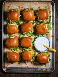 Buffalo Chicken Sliders on a lined sheet tray with a small round dish of blue cheese sauce on the side.