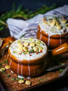 Two crocks of New England Clam Chowder on a tray with bread, oyster crackers and herbs scattered around.
