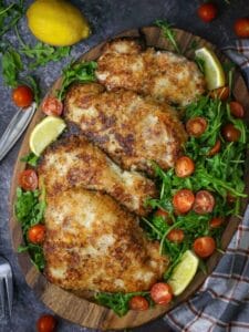 Pork Milanese with Arugula and Tomatoes on an oval wood board with lemon and arugula scattered around.