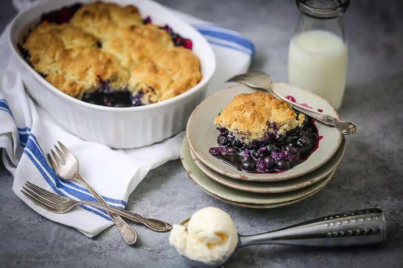 Blueberry Cobbler in the baking dish and on a small serving plate with ice cream scooped on the table.