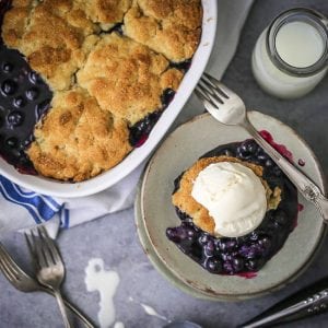 A serving of Blueberry Cobbler topped with vanilla ice cream next to the cobbler dish.
