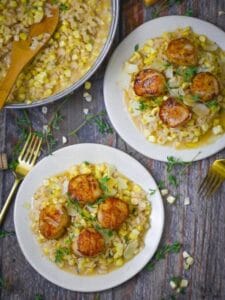 Two plates of seared scallops over creamy fresh corn risotto next to risotto in the pan and a wedge of parmesan cheese.