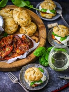 Tomato "Shortcake" with Whipped Ricotta ingredients on a round wood board with basil scattered around and assembled shortcakes and wine on the table.