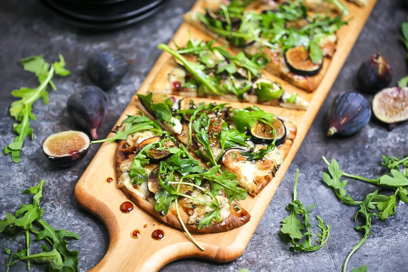 Close up of fig, blue cheese, and arugula flatbread sliced and served on a wooden cutting board and drizzled with honey balsamic vinaigrette.